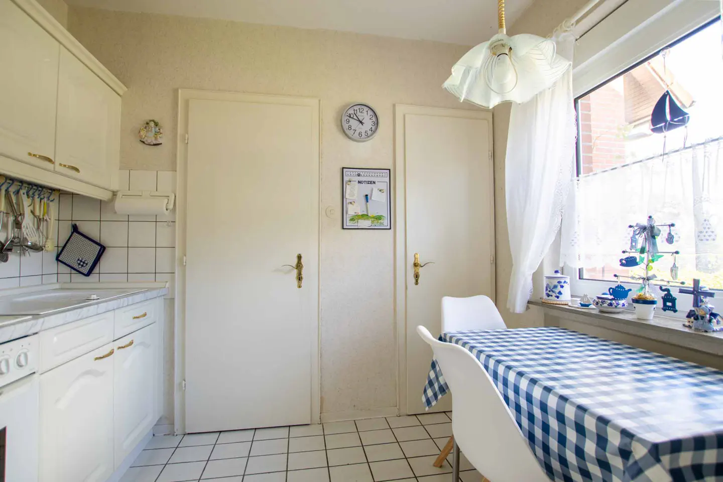 Bright kitchen with white cabinets, tile floor, and a table covered with a blue and white checkered tablecloth. Two white doors and a window with white curtains.