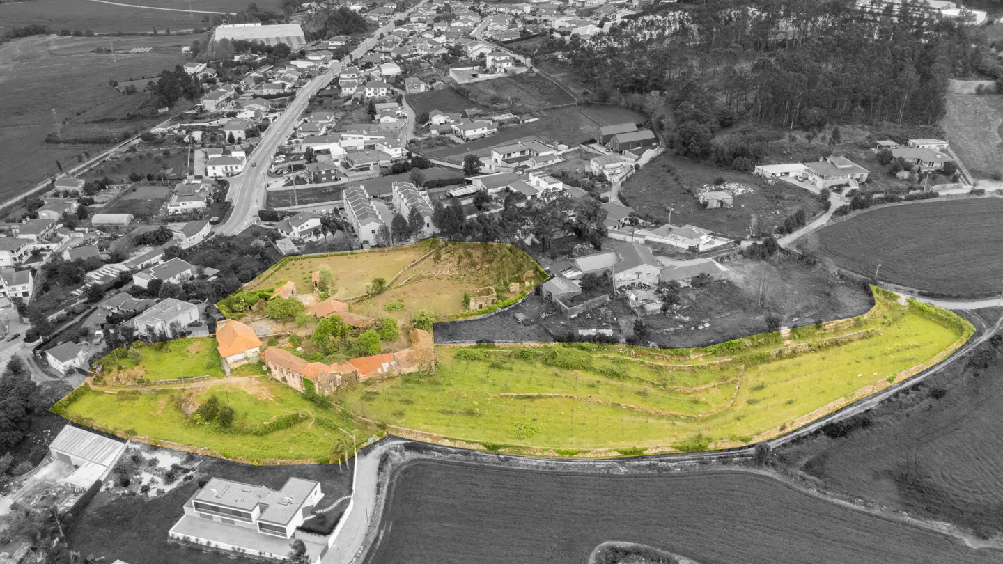 Aerial view of a property outlined in yellow, featuring a stone building with a red tile roof and green grass, set against a grayscale town.