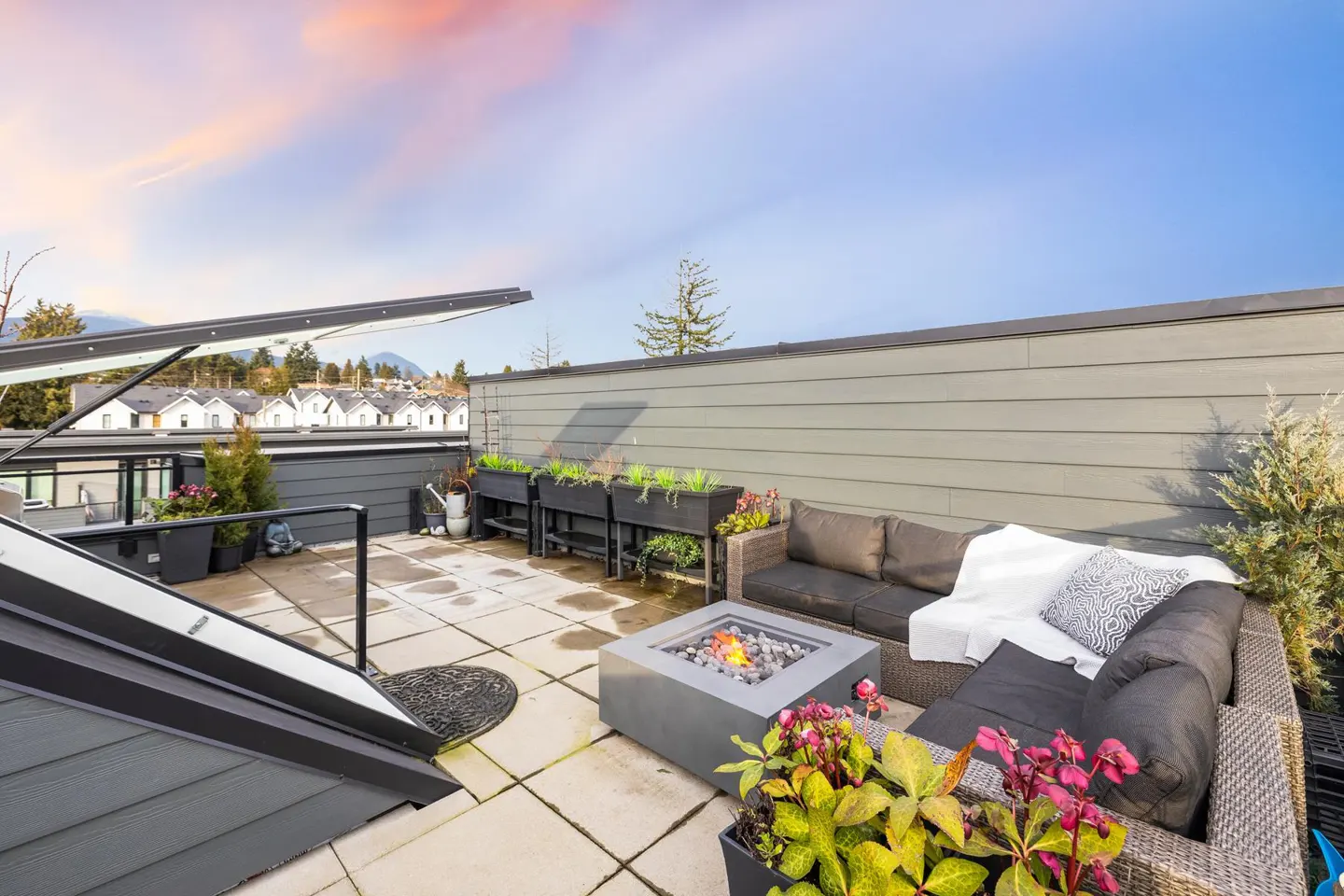 Rooftop patio with a fire pit, grey sectional sofa, potted plants, and a sunset sky over a suburban neighborhood.