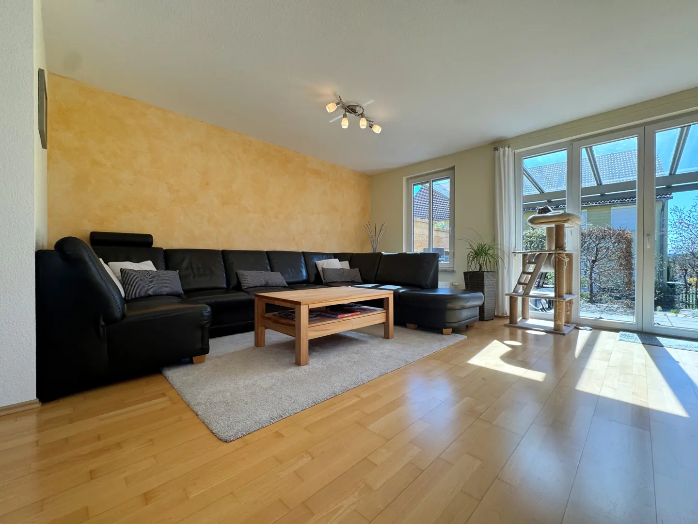 Bright living room with wood floors, a large black leather sectional, and a wooden coffee table on a gray rug. A cat tree stands near glass doors.