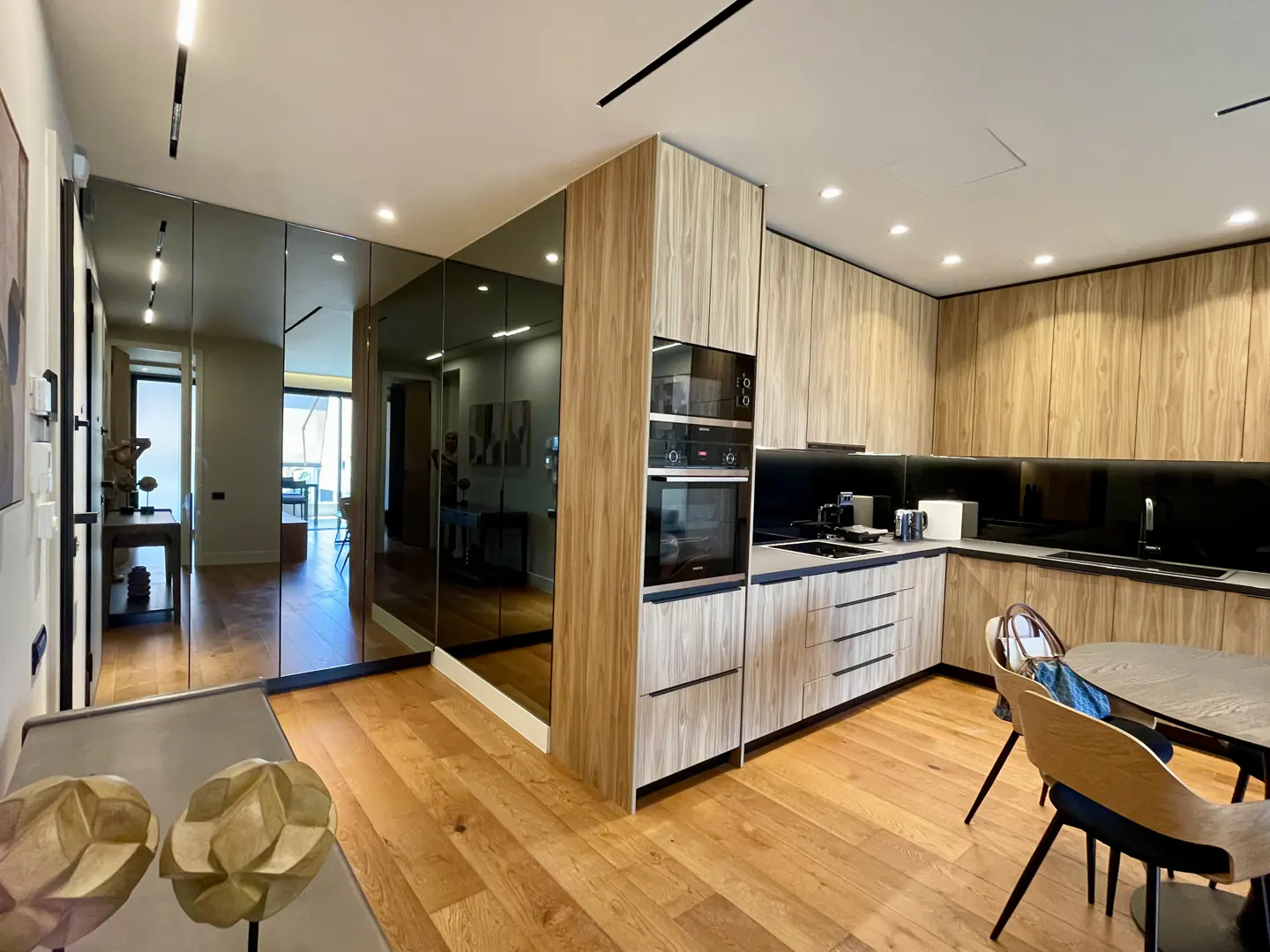 Modern kitchen with wood cabinets, black countertops, and stainless steel appliances. Mirrored wall reflects hallway. Hardwood floors throughout.