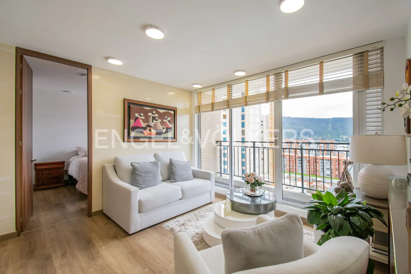 Bright living room with white sofa, round tables, and balcony overlooking city. Bedroom visible through open door.