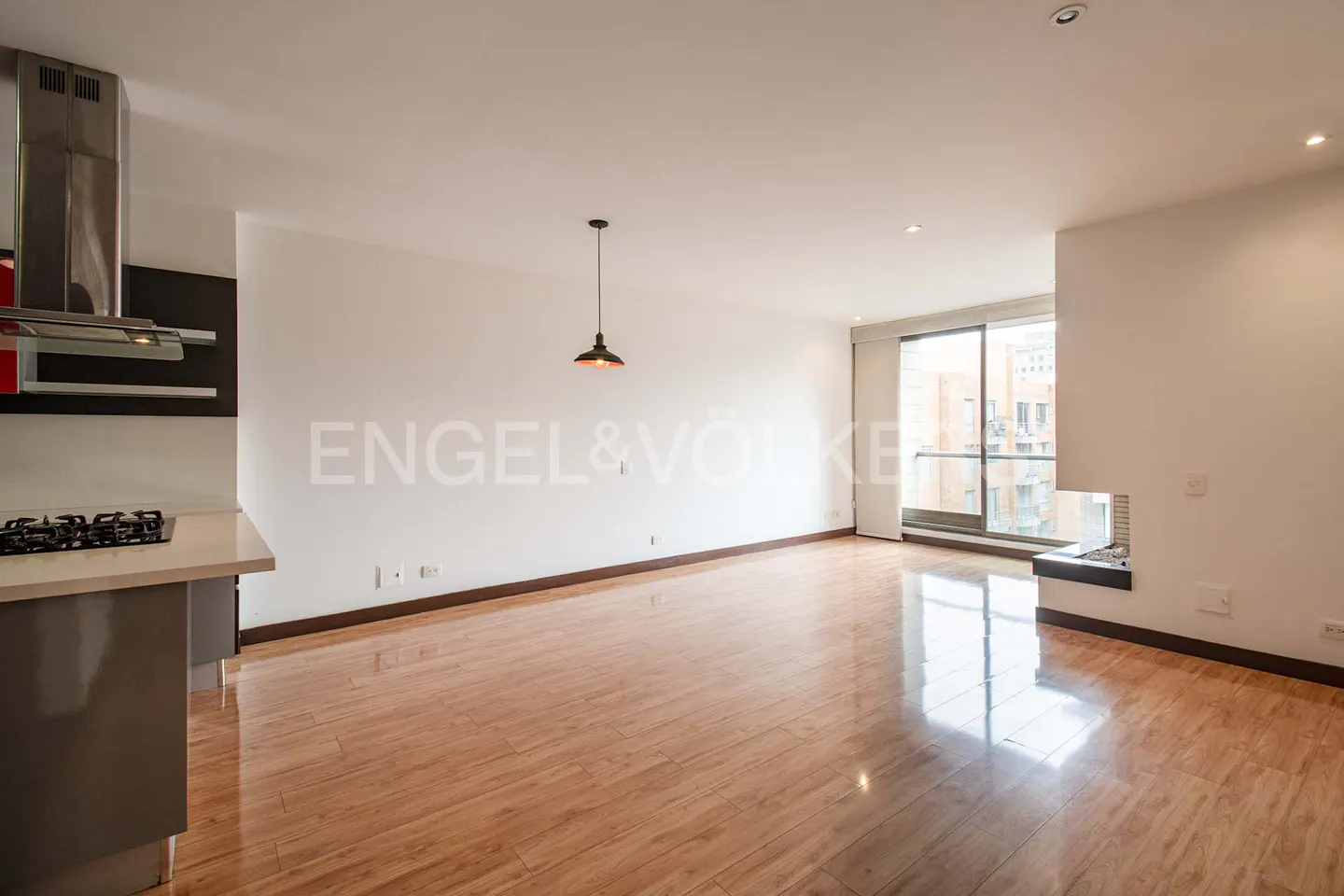Bright, empty apartment with wood floors, white walls, and a modern kitchen area. A pendant light hangs in the center of the room. Large windows offer a city view.