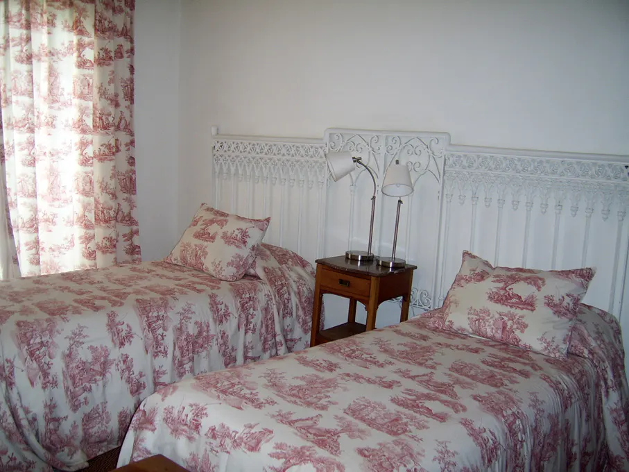 Bedroom with two twin beds, white iron headboards, red toile bedding, matching curtains, and a wooden nightstand with lamps.