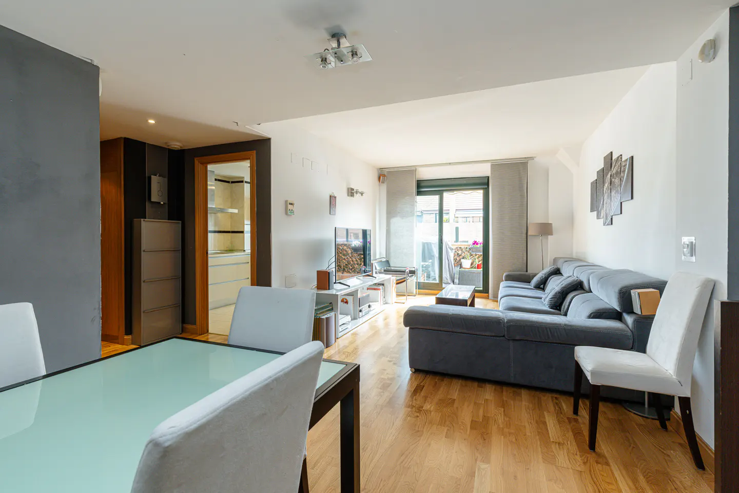 Bright living room with wood floors, gray sofa, and glass dining table with white chairs. Balcony with flowers visible.