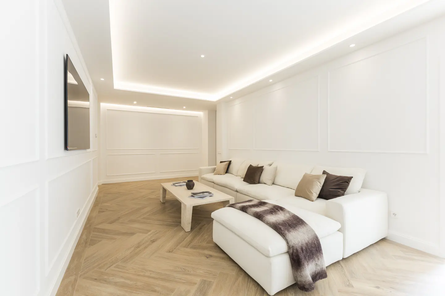 Bright living room with white walls, herringbone wood floors, and recessed lighting. A white sectional sofa with brown pillows faces a white coffee table. A TV hangs on the wall.