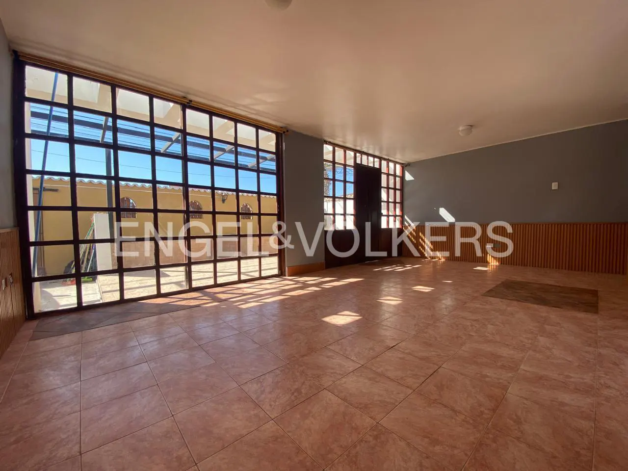 An empty room with terracotta tile flooring, large windows, and the Engel & Volkers logo.