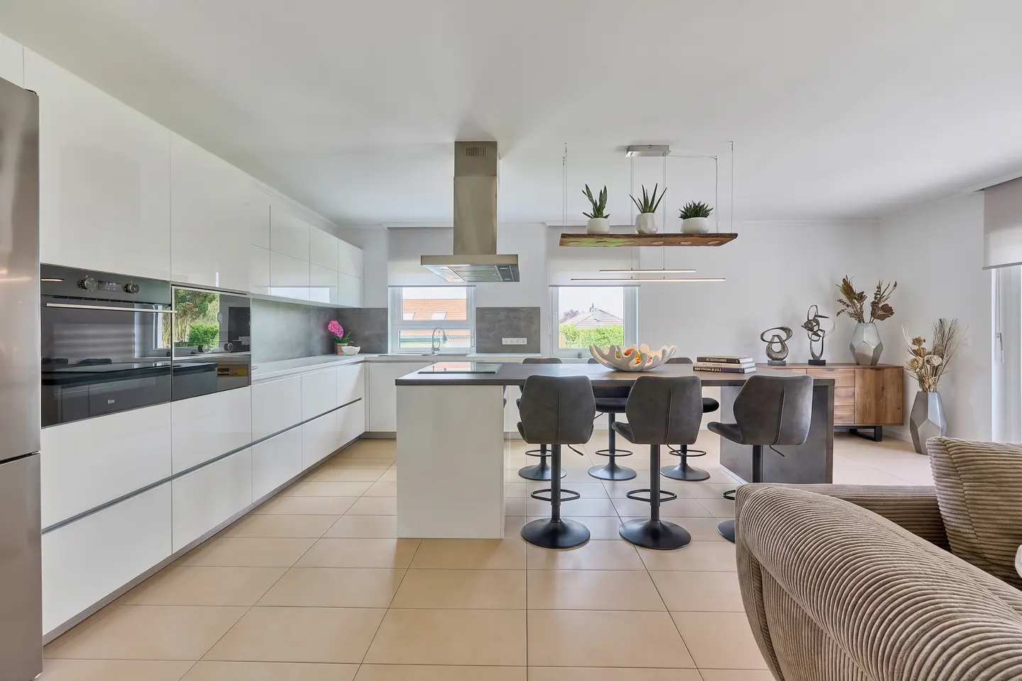 Bright, modern kitchen with white cabinets, stainless steel appliances, and a central island with gray stools. A wooden shelf with plants hangs above.
