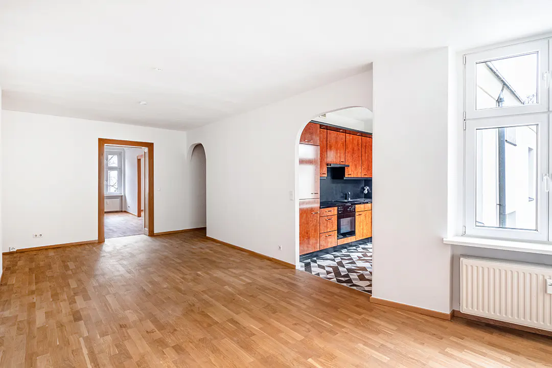 An interior shot of a bright, empty apartment with hardwood floors and white walls, showcasing the kitchen through an arched doorway.