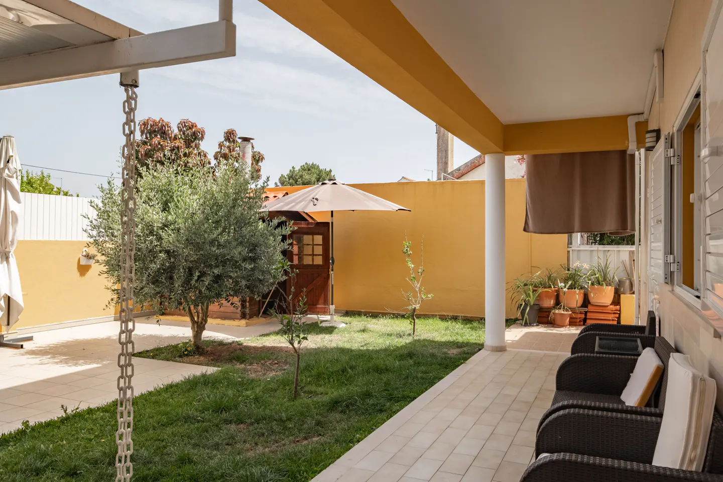 Outdoor patio with wicker chairs and cushions, overlooking a green lawn with trees and a small wooden shed. Yellow walls and a beige umbrella are visible.