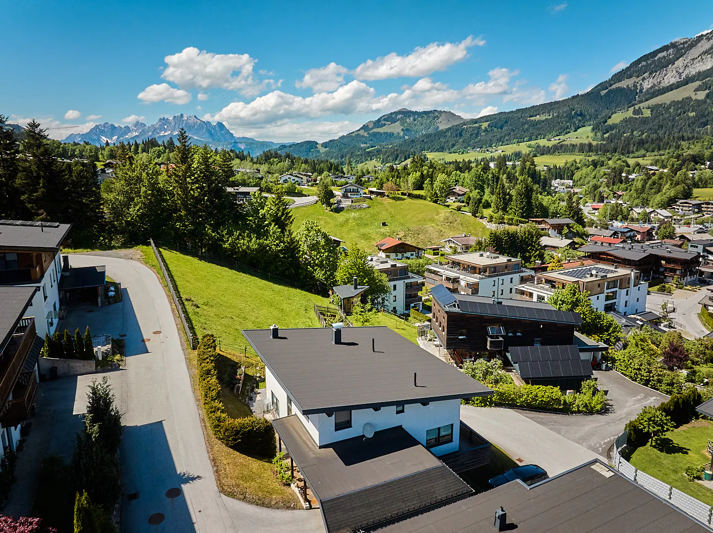 Scenic aerial view of a village nestled in a green valley, with mountains and a blue sky with clouds in the background.