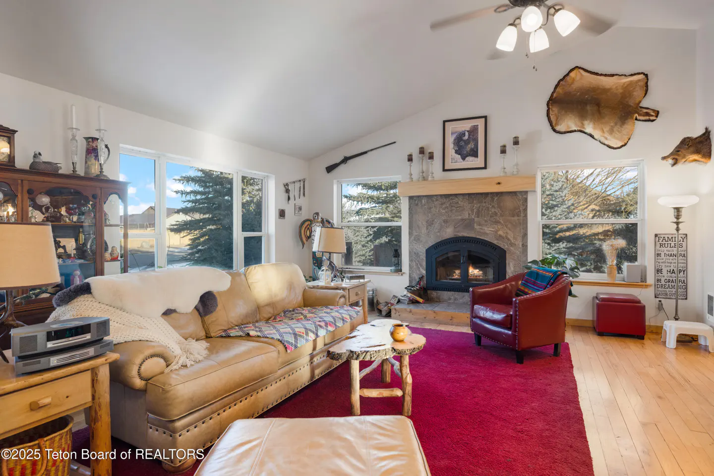 Living room with a tan leather sofa, red rug, stone fireplace, and large windows with a view of trees.
