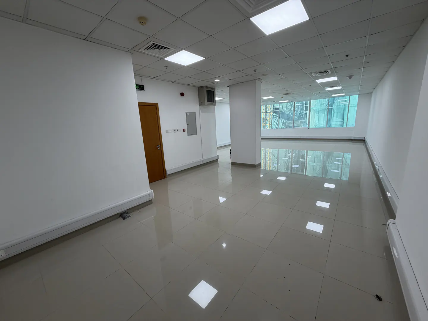 Bright, empty office space with white walls, tile floors, and large windows. A brown door is on the left wall.
