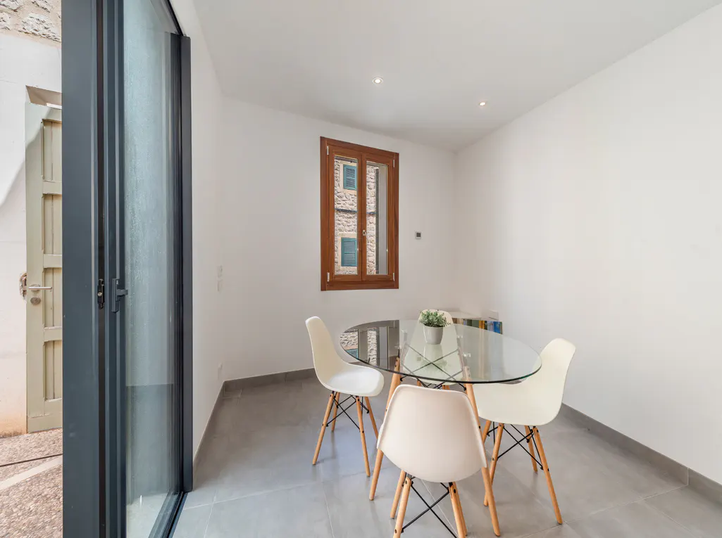 Bright dining room with white walls, gray tile floor, and a glass table surrounded by four white chairs. A window and a sliding glass door provide natural light.
