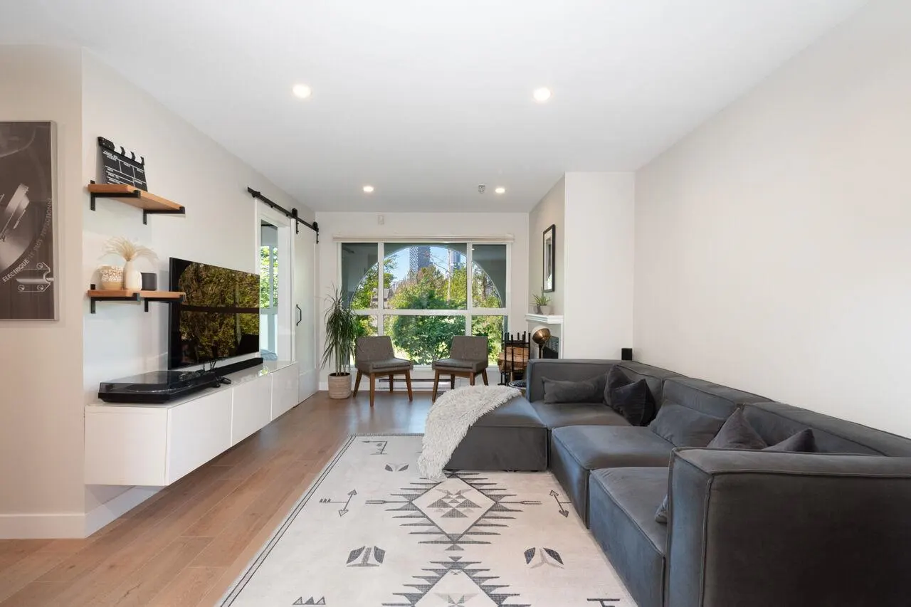 Living room with gray sectional sofa, patterned rug, TV on white console, shelves, and arched window with chairs.