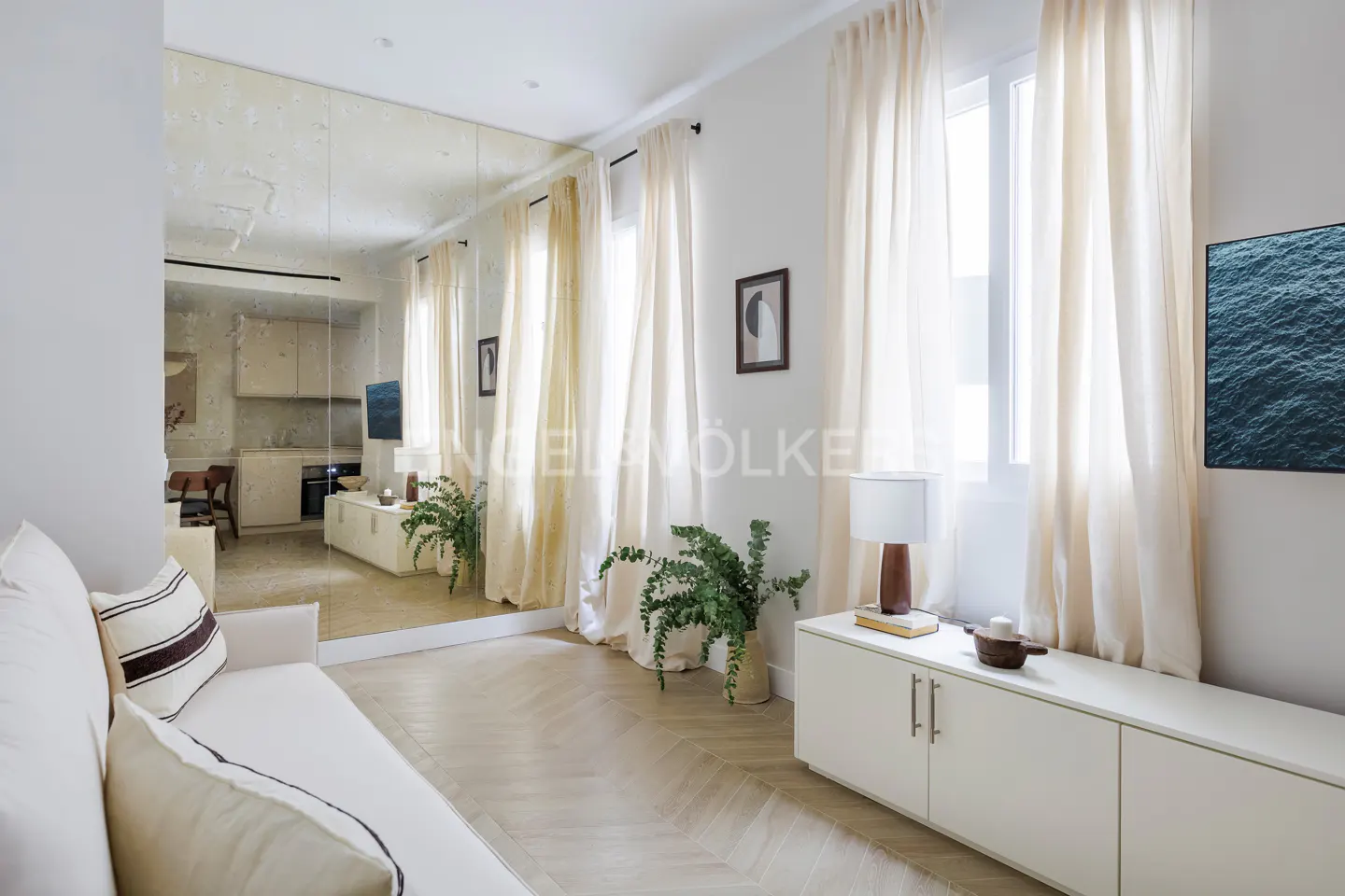 Living room with a white sofa, herringbone floor, and a large mirror reflecting the kitchen. Cream-colored curtains frame a window.