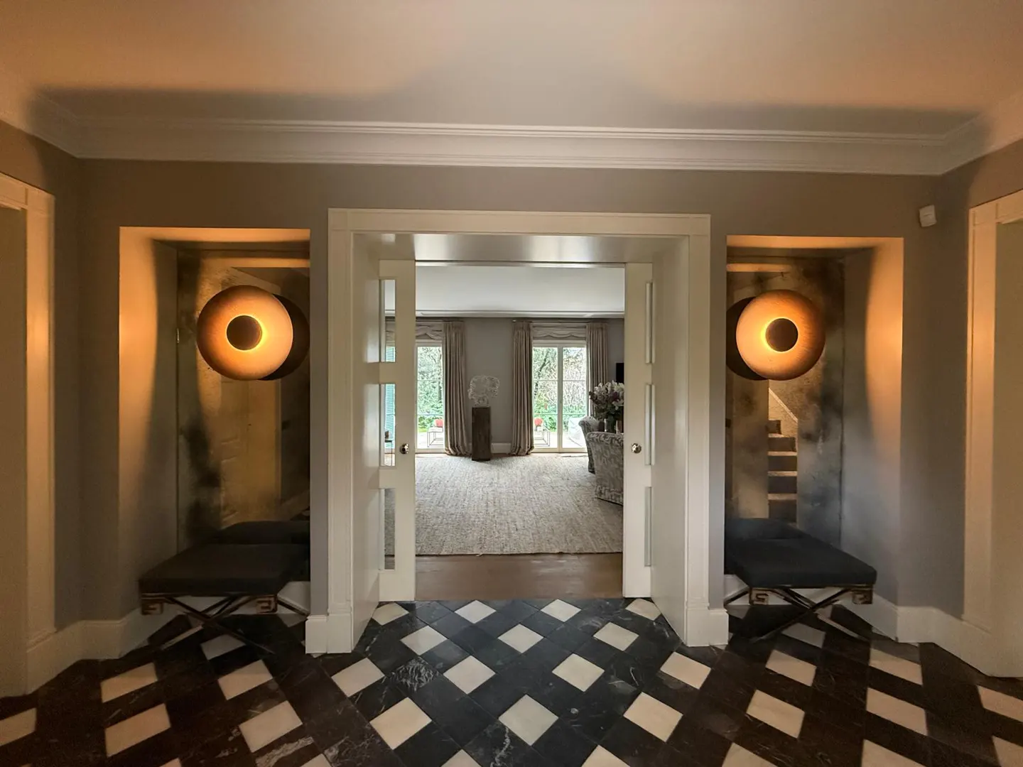Entryway with black and white checkered floor, two dark benches, wall sconces, and a view into a bright living room.