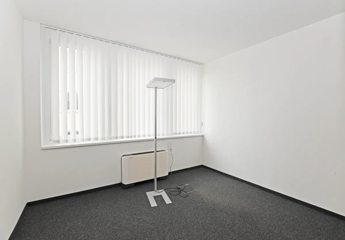 Empty office space with gray carpet, white walls, and a window with white blinds. A standing lamp and a heater are near the window.