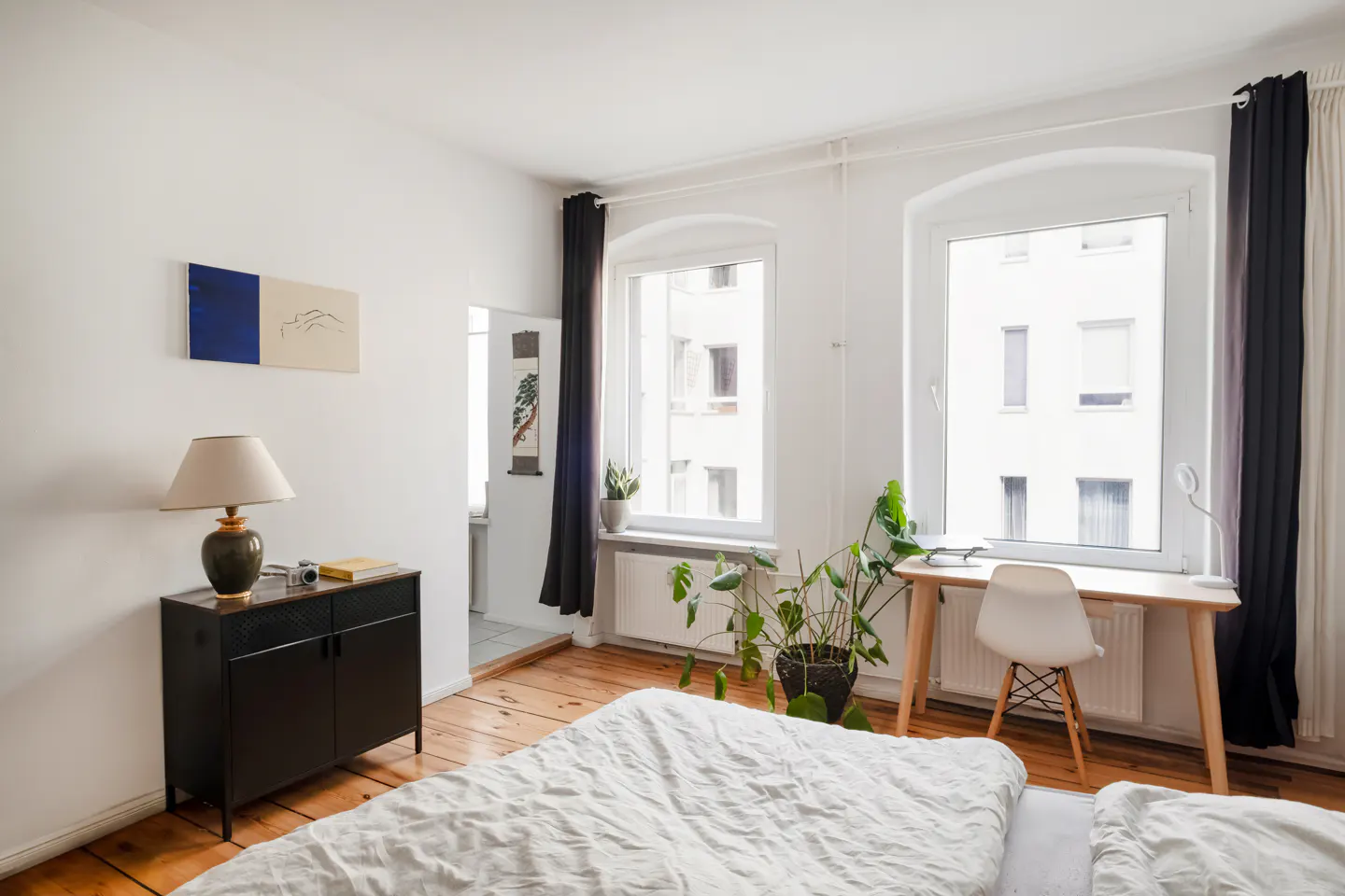 Bright bedroom with white walls, wood floors, and two windows. A bed with white linens is in the foreground. A desk, chair, and black cabinet are also visible.