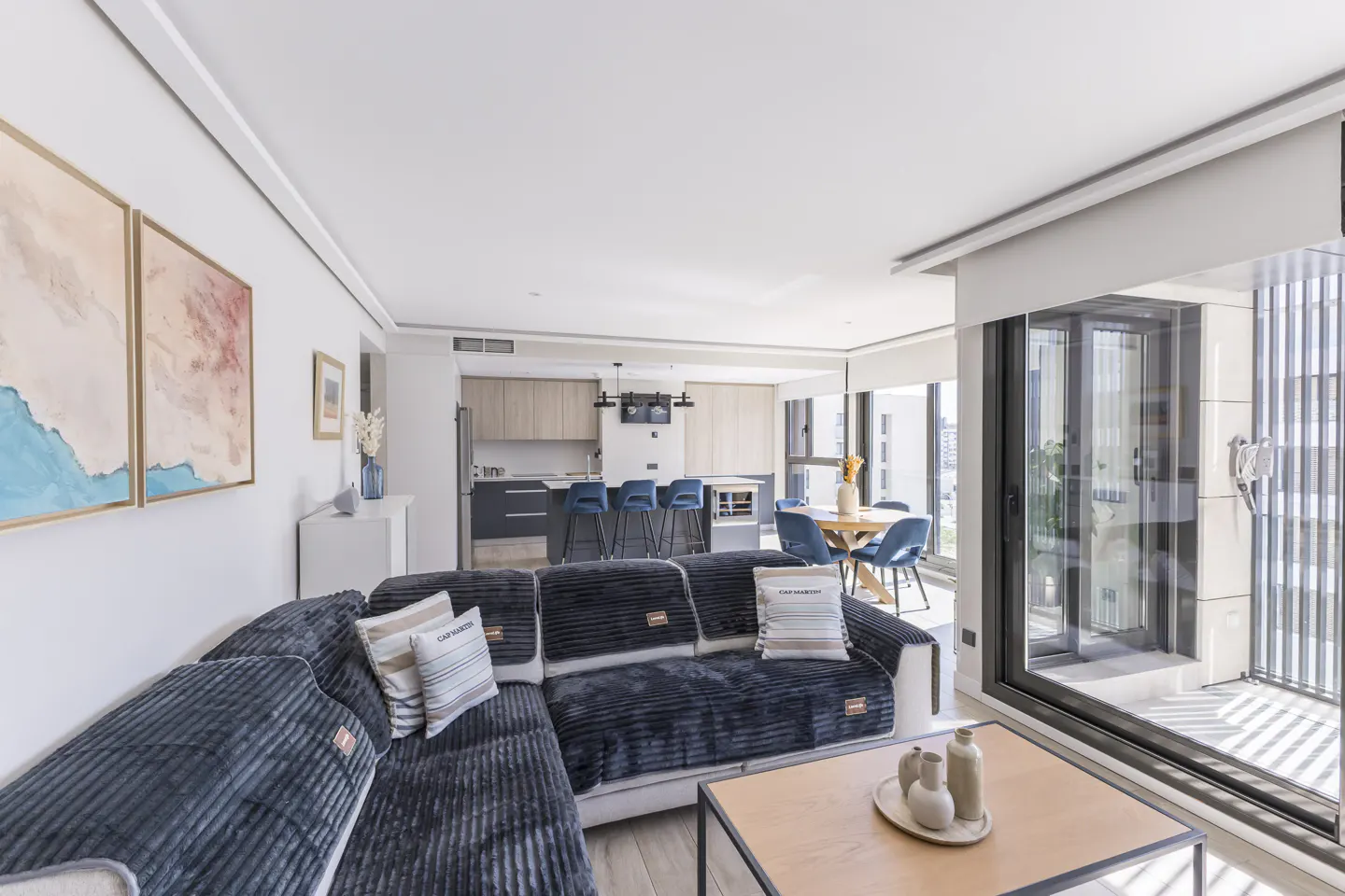 Open-concept living space with a blue corduroy sofa, wood coffee table, and kitchen with blue bar stools. Large windows offer city views.