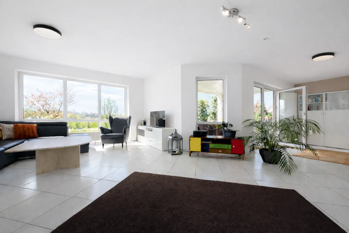 Bright living room with white walls, tile floors, and large windows. Features a black sofa, gray armchair, and colorful cabinet.