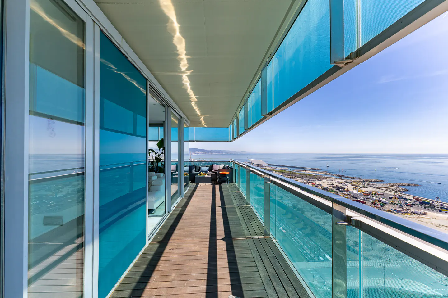 A long balcony with blue glass panels and a wooden floor overlooks the ocean and a city.
