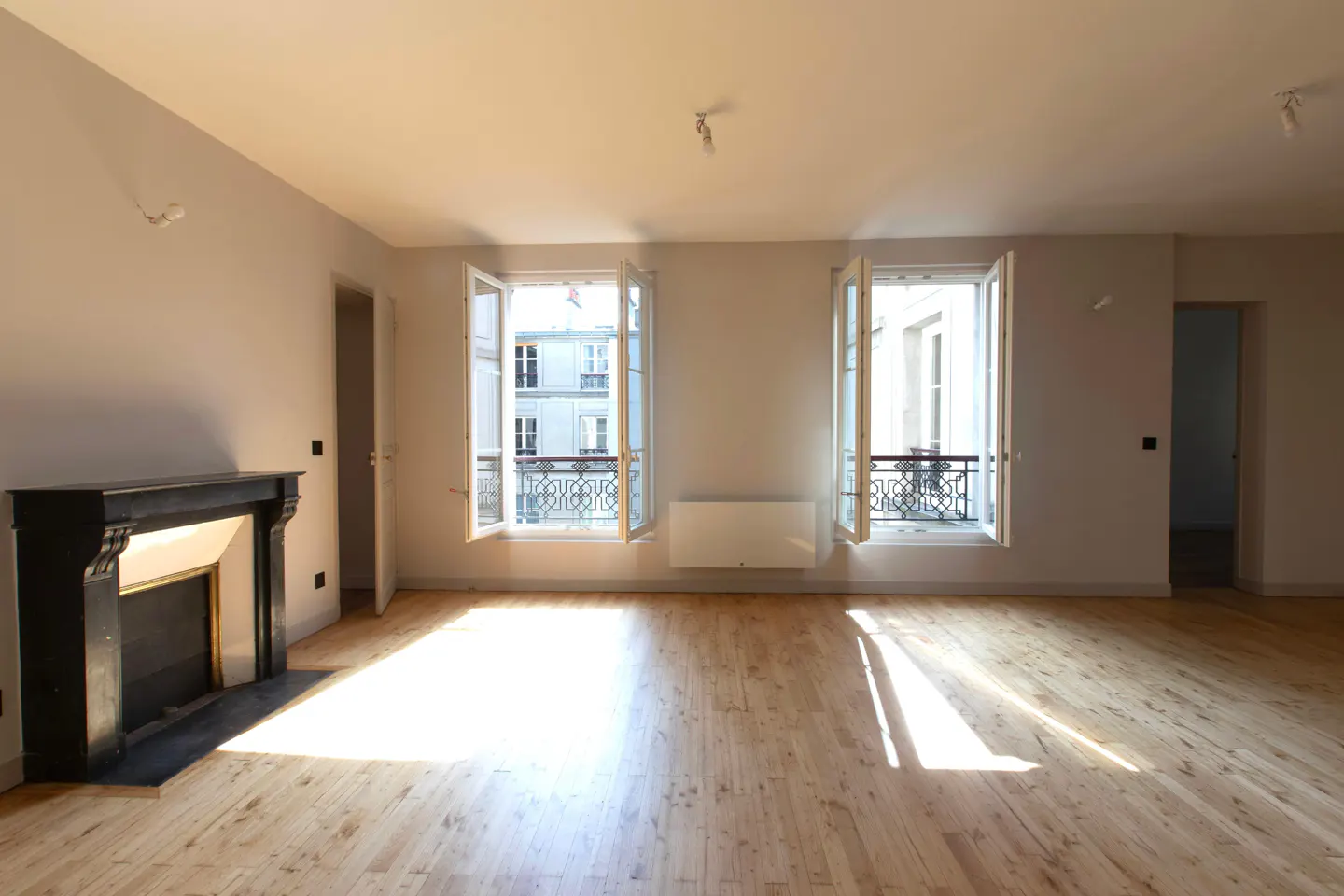 Bright, empty room with hardwood floors, white walls, and a black fireplace. Two open windows reveal a building with balconies.
