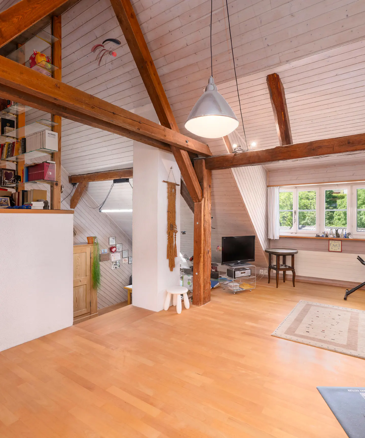 Attic room with wooden beams, white walls, and light wood floors. A TV, table, and rug furnish the space. Windows offer a view of greenery.
