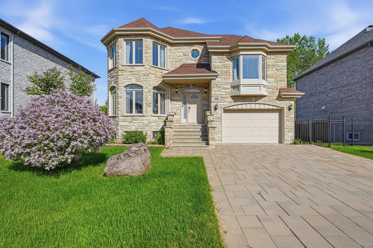 Two-story stone house with a brown roof, paved driveway, and a large blooming lilac bush in the front yard.