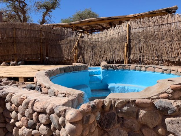 Outdoor pool with stone border and steps, surrounded by a reed fence and a bamboo roof structure. Blue sky in the background.