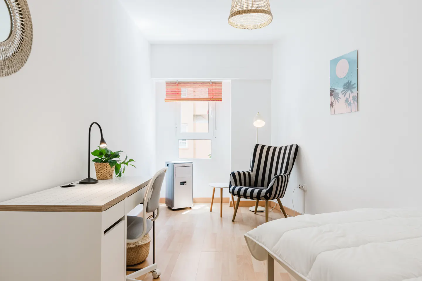Bright bedroom with white walls, wood floor, desk, striped chair, and bed. A woven lamp hangs from the ceiling.