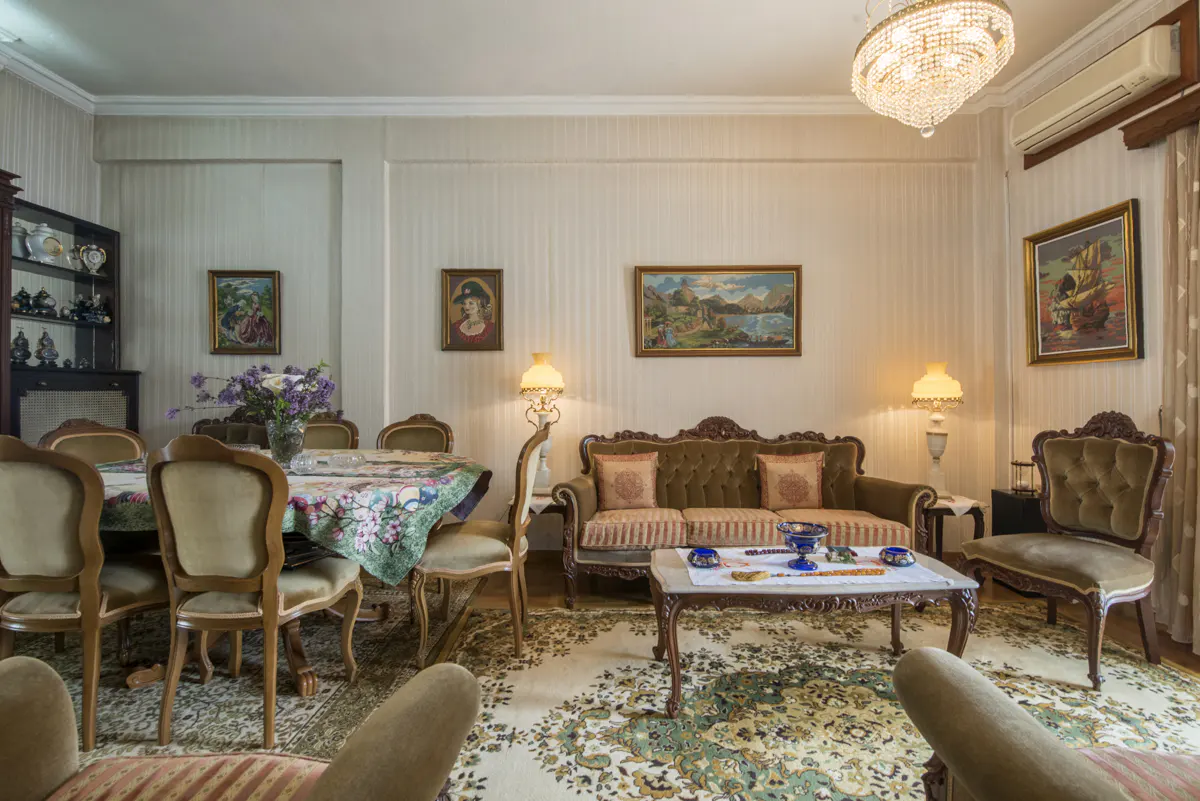 Ornate living and dining room with floral rug, olive sofa, and dining table with floral tablecloth. Paintings adorn the walls.