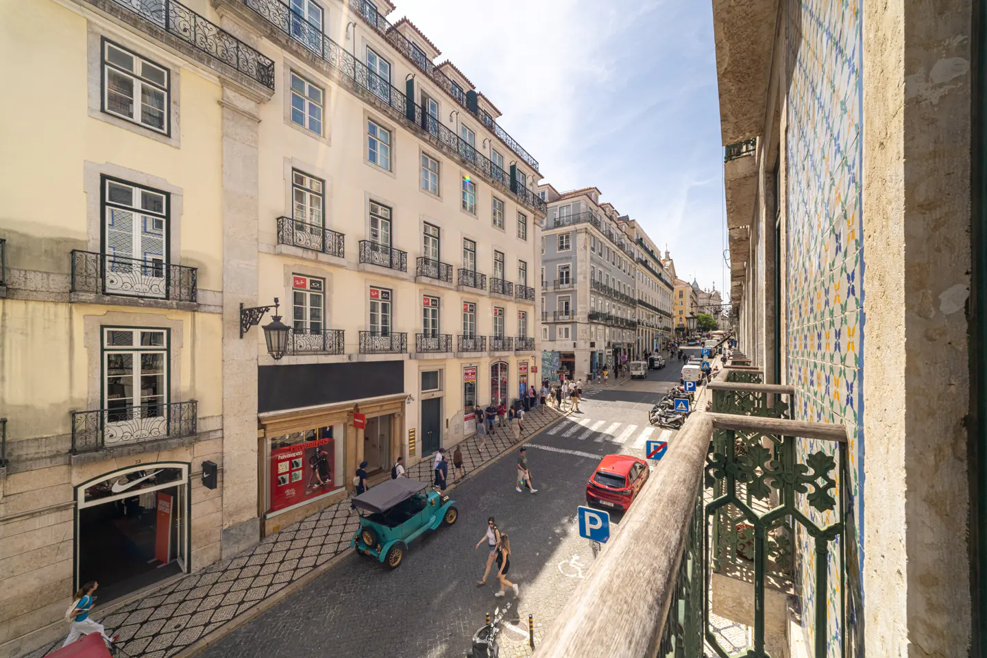 View from a balcony overlooking a Lisbon street with cars, pedestrians, and buildings. The balcony has decorative tiles and iron railings.