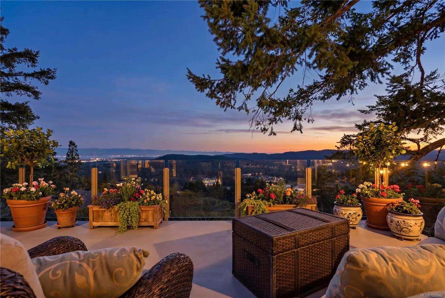 Outdoor patio with wicker furniture, potted plants, and a sunset view over a city skyline. Glass railing provides safety.