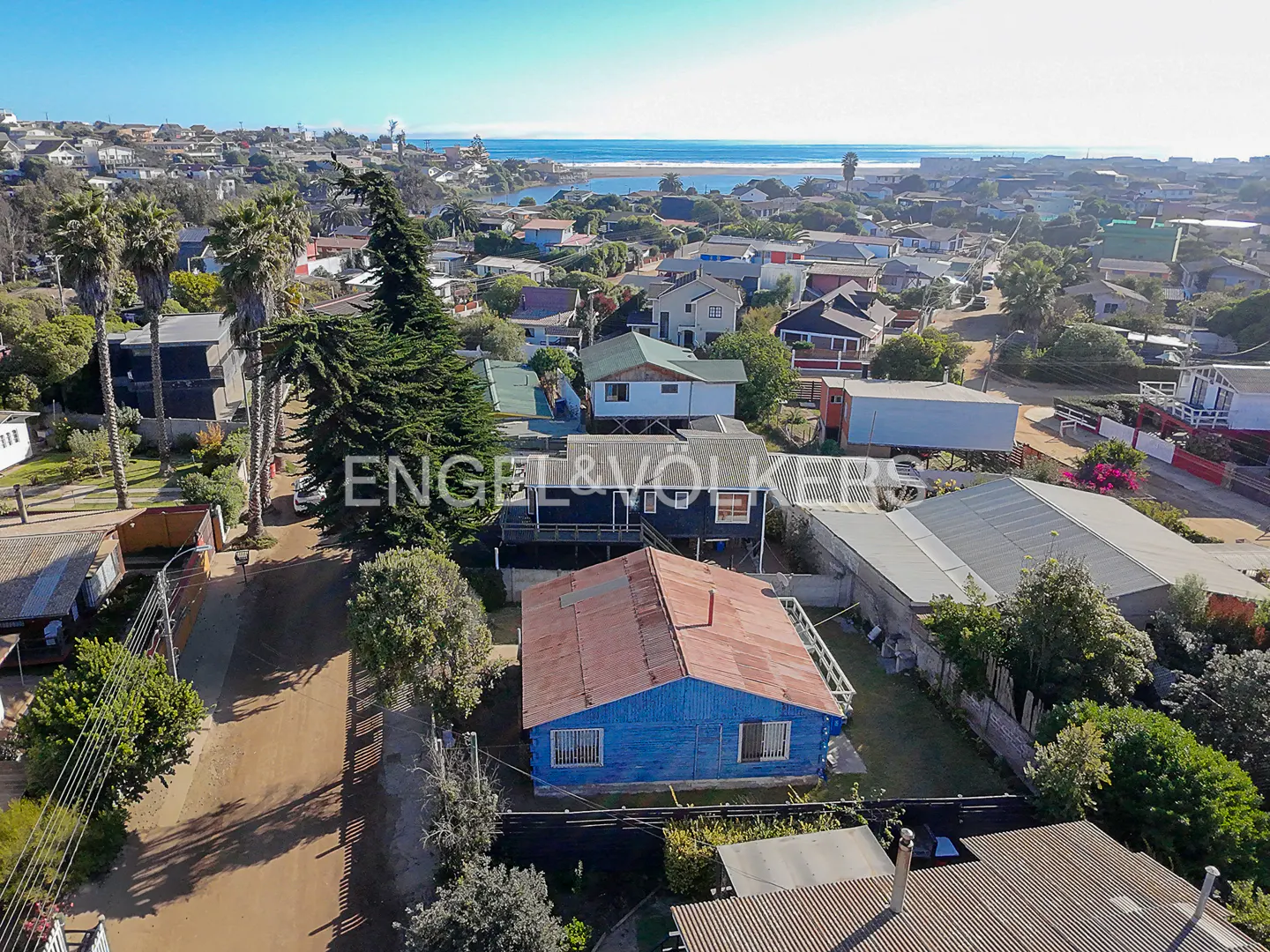 Aerial view of a coastal town with colorful houses, palm trees, and the ocean in the background under a clear blue sky.