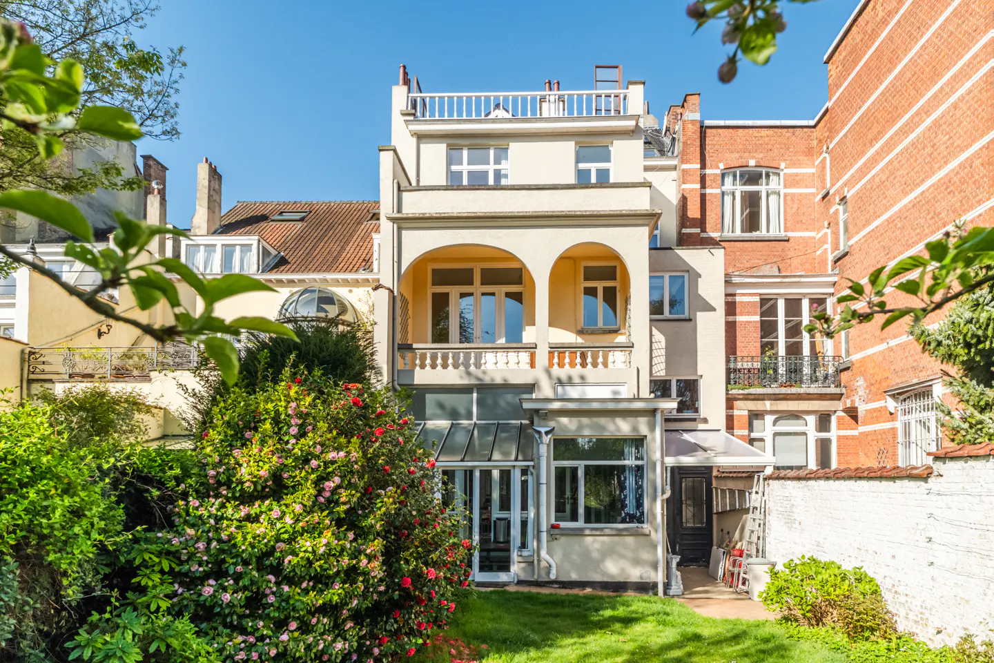 Back view of a cream-colored three-story house with a rooftop deck, surrounded by green bushes and grass.