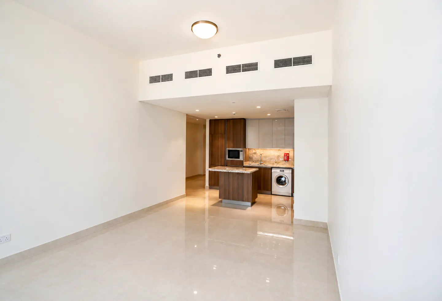 An empty, bright apartment with white walls and tile floors. An open kitchen with brown cabinets, a microwave, and a washing machine is visible.