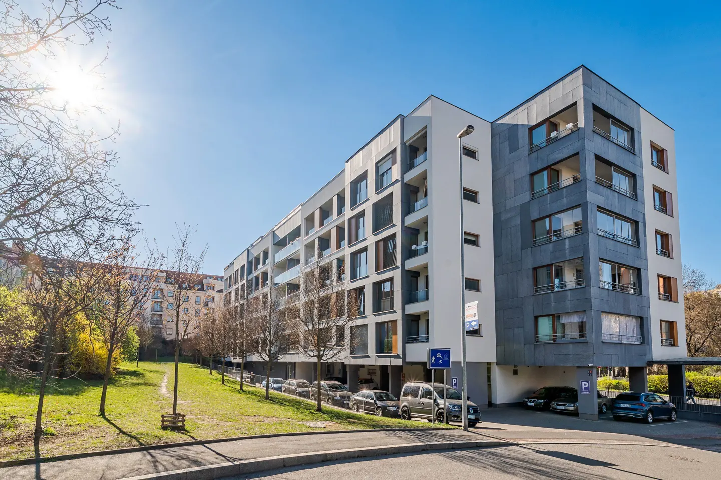 Modern apartment building with gray and white facade, balconies, and parking. Sunny day with blue sky.