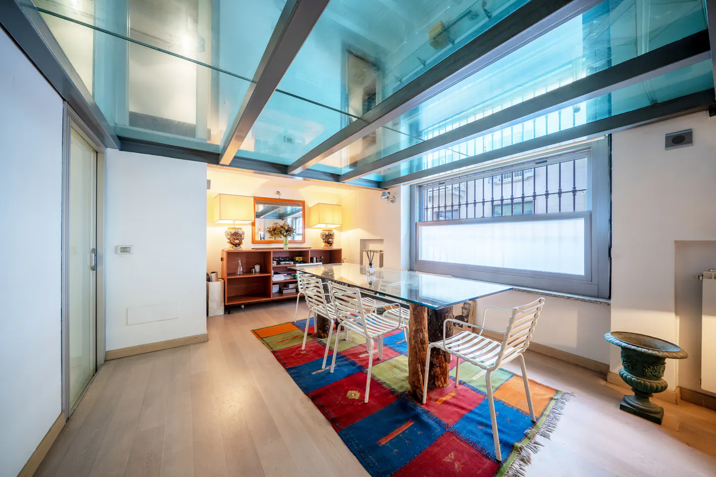 Bright dining room with glass ceiling, wood floors, and colorful rug. A glass table with tree trunk legs is surrounded by white chairs.