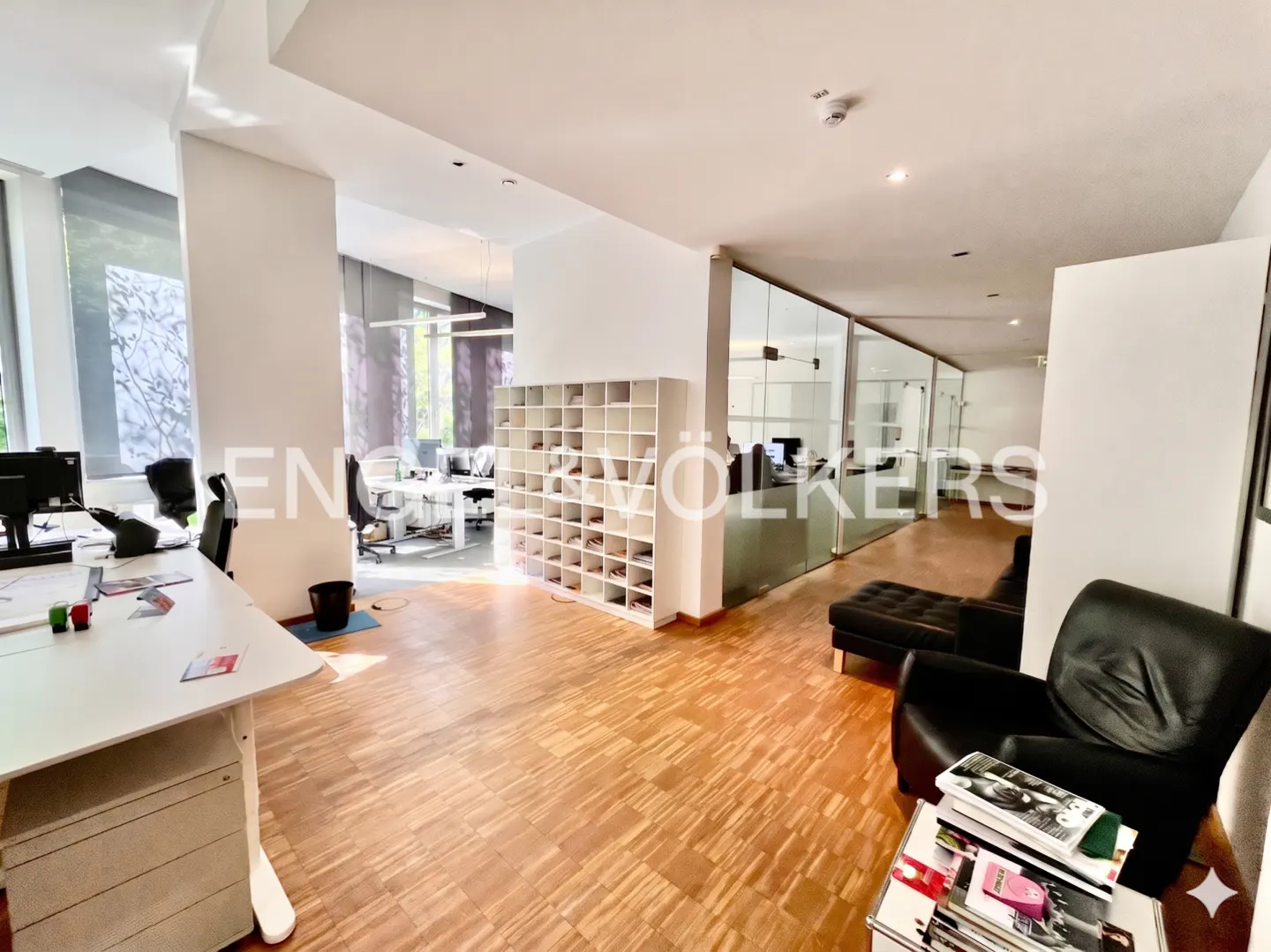 Office space with wood floors, white walls, desks, chairs, and glass-walled offices. A black leather chair and ottoman are in the foreground.