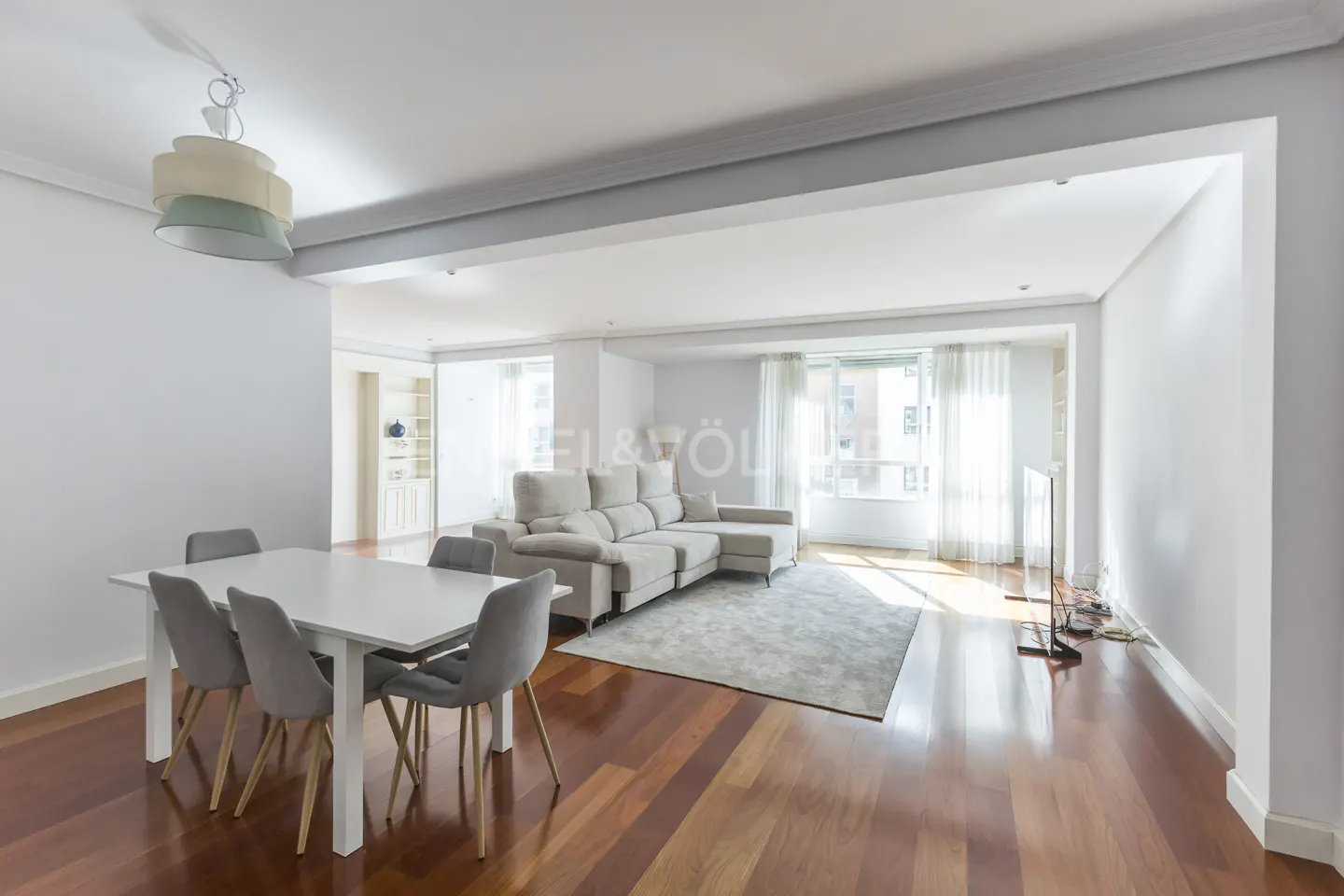 Bright, open-plan living and dining area with hardwood floors, white walls, and a large beige sectional sofa. A white dining table with gray chairs sits in the foreground.