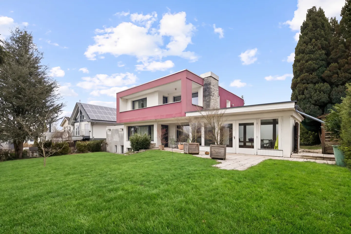 Two-story house with a red facade, stone chimney, and a green lawn under a blue sky with white clouds.
