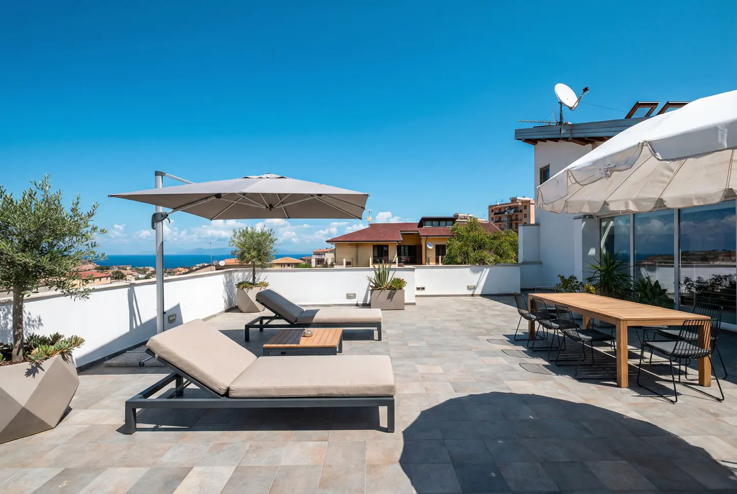Rooftop patio with lounge chairs, dining table, and umbrellas. Ocean view in the background under a clear blue sky.