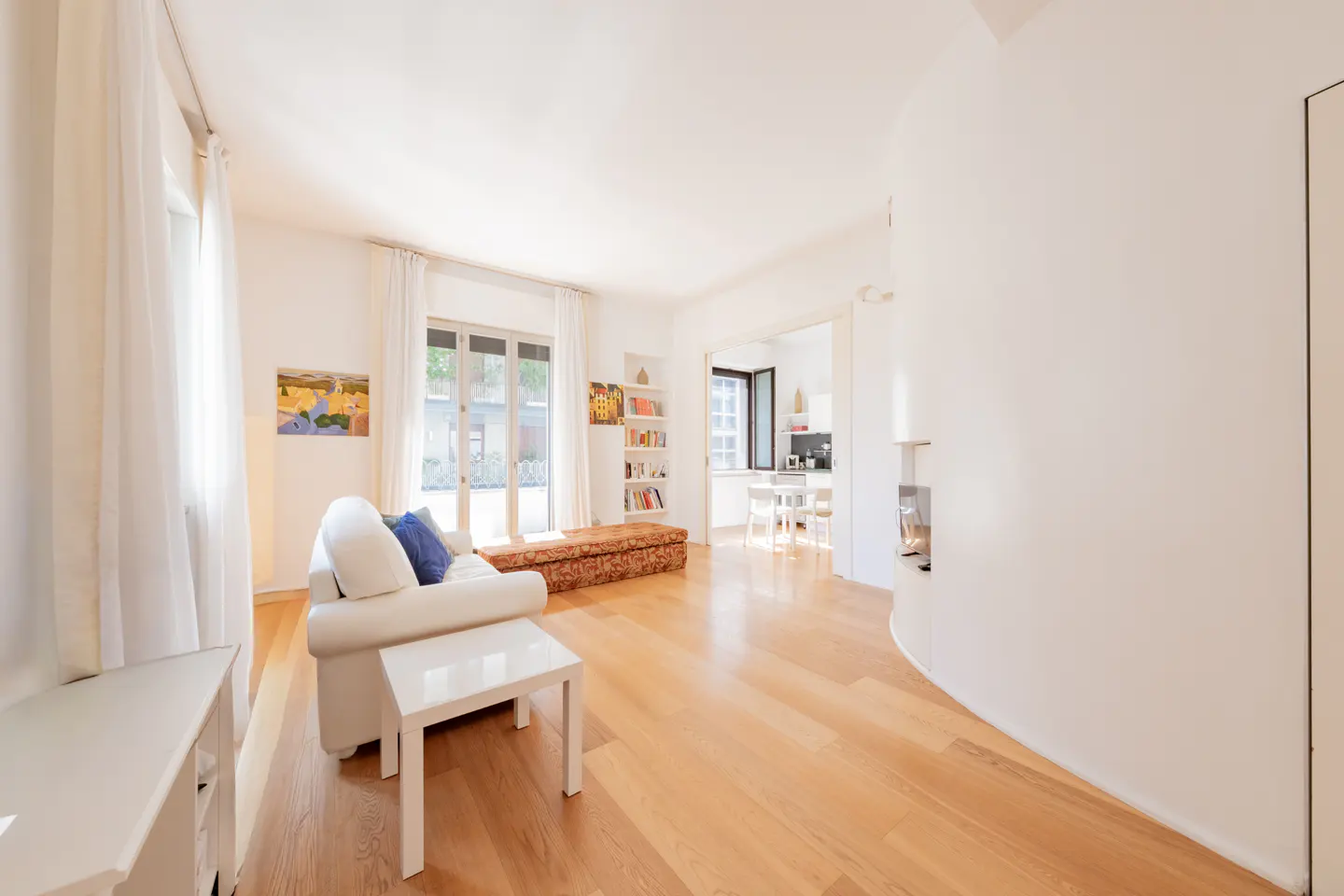 Bright living room with hardwood floors, white walls, and a white sofa with blue pillows. A small white table sits in front of the sofa. Bookshelves and a kitchen are visible in the background.