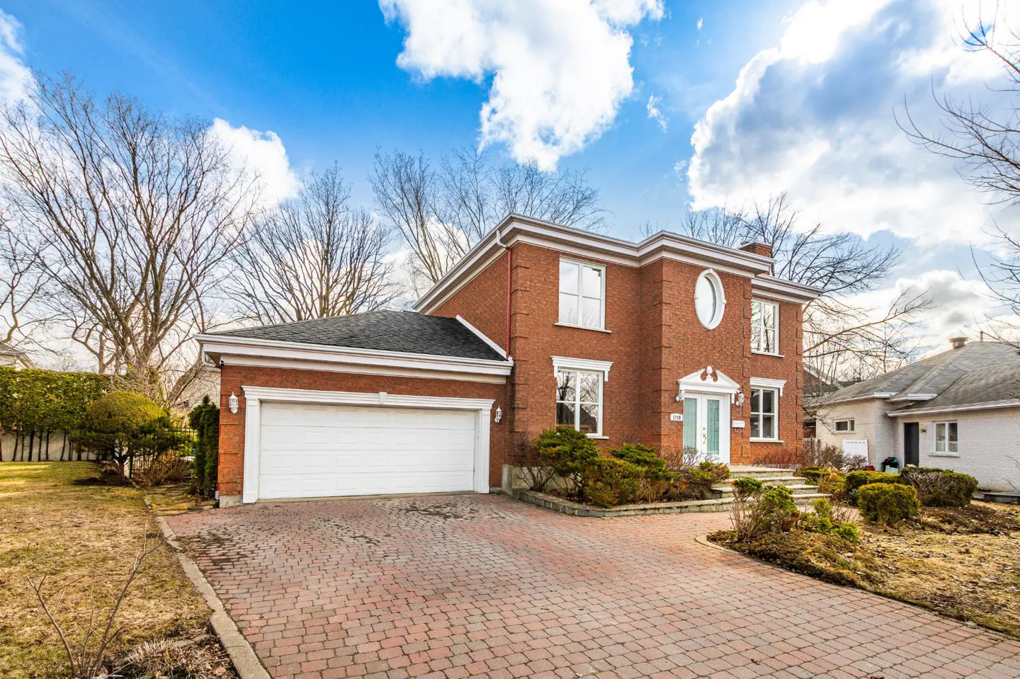 Two-story red brick house with a white garage door and a brick driveway under a blue sky.