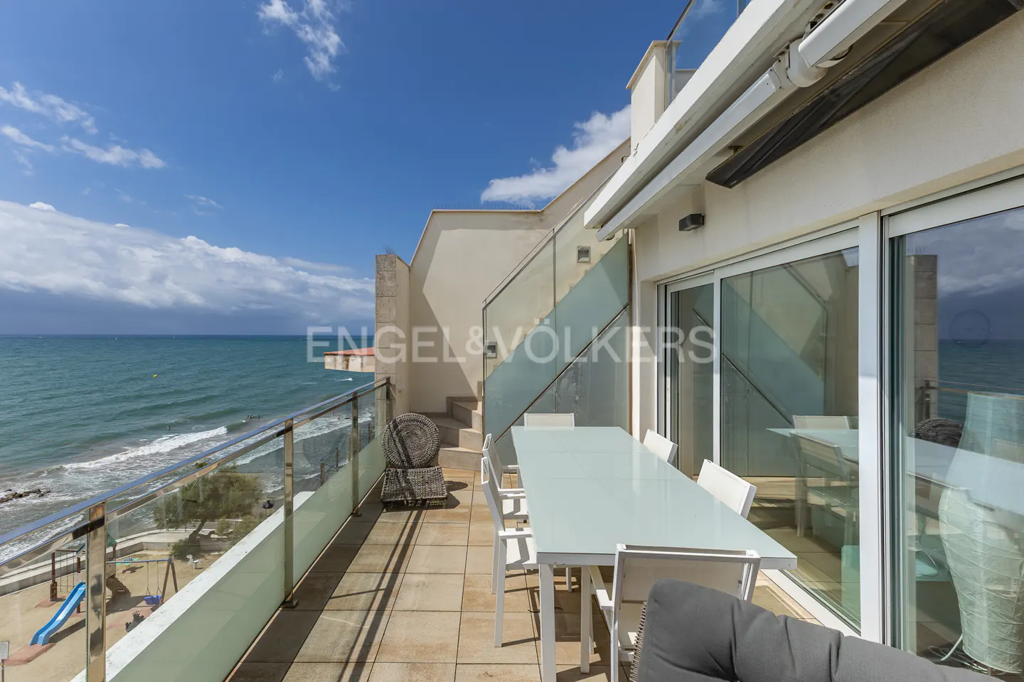 Balcony with ocean view. White table and chairs on a tiled patio. Glass railings overlook the beach and blue sea.