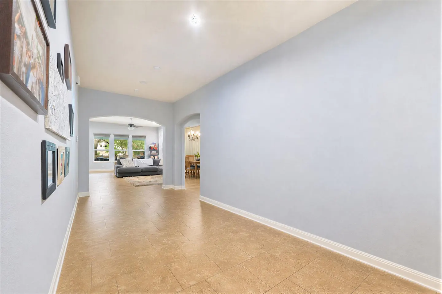Hallway with light blue walls, tan tile flooring, and framed pictures on the left wall. Arched doorways lead to a living room and dining area.