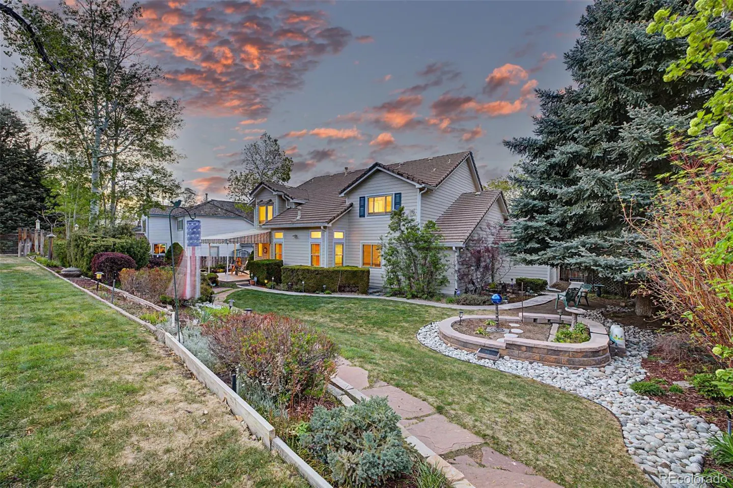 Exterior view of a two-story house with a landscaped yard at dusk. The house is light-colored with a brown roof and blue shutters.