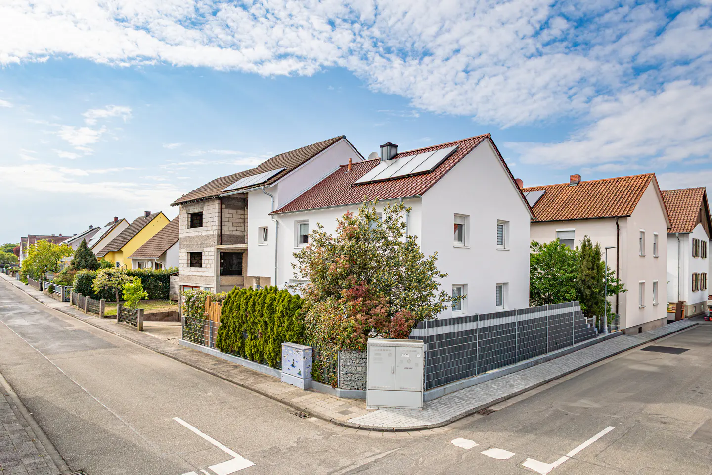 Street view of houses with red and brown roofs, some with solar panels, under a cloudy blue sky.