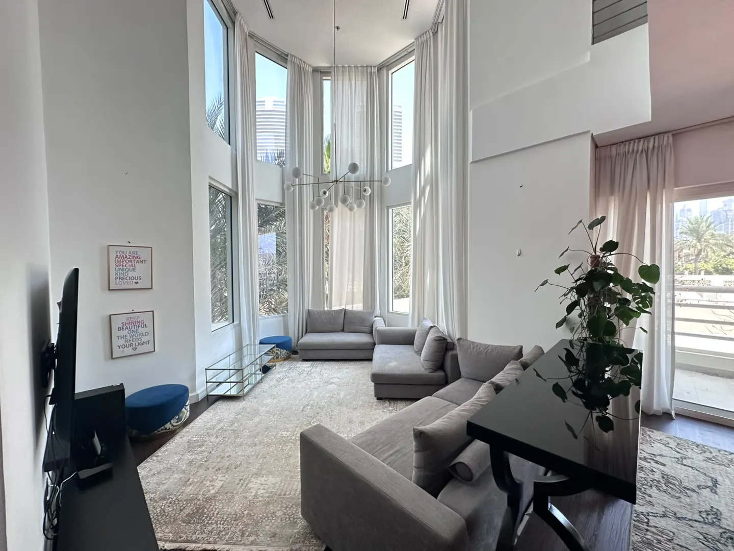 Bright living room with high ceilings, floor-to-ceiling white curtains, grey sectional sofas, and a modern chandelier.